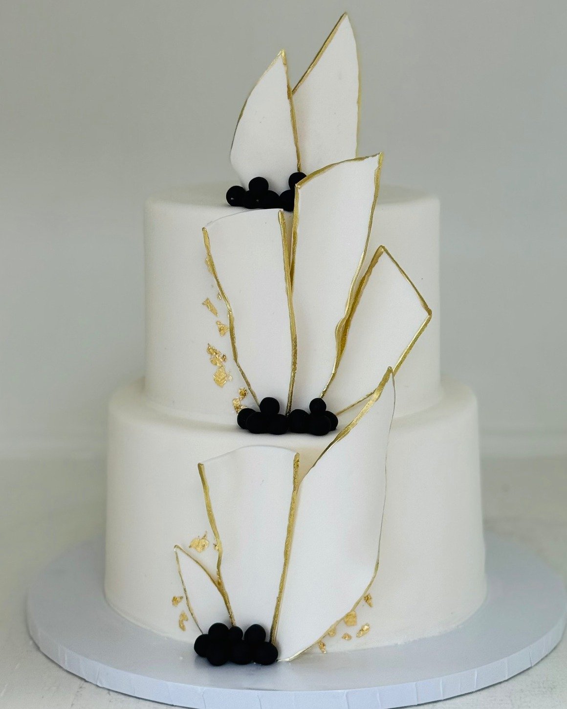 A modern two-tier white wedding cake with gold-edged fondant shards and gold leaf, a sculptural 2026 wedding cake by Sweet Escape Cake Company in Indianapolis.
