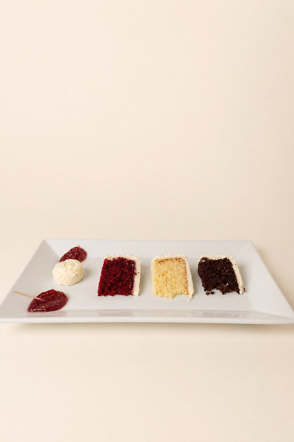 A formal cake tasting featuring slices of red velvet, vanilla, and chocolate cakes, accompanied by cream and berry jam, presented on white r