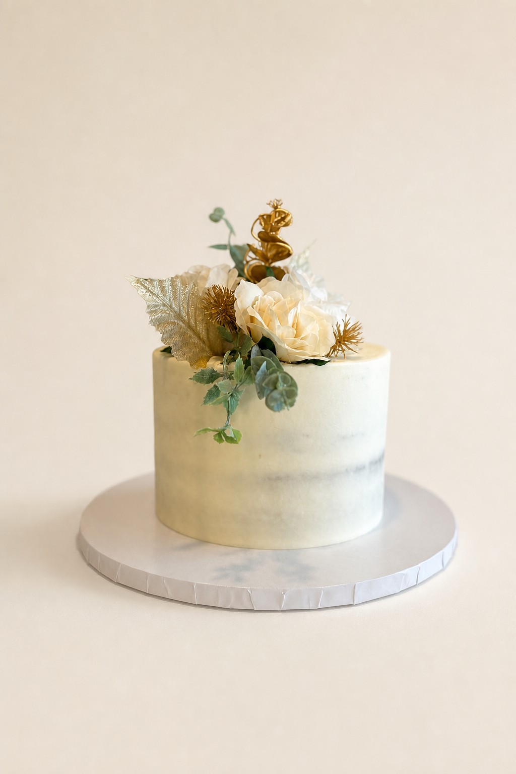 A single-tier round naked cake decorated with cream-colored flowers, shimmering gold leaves, green foliage, and a delicate gold topper, perf