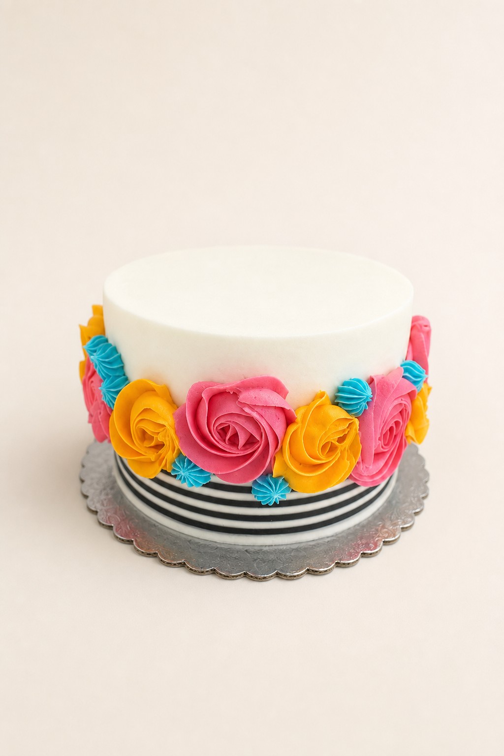 A festive single-tier round white buttercream cake adorned with a vibrant band of piped hot pink, yellow, and turquoise buttercream flowers 