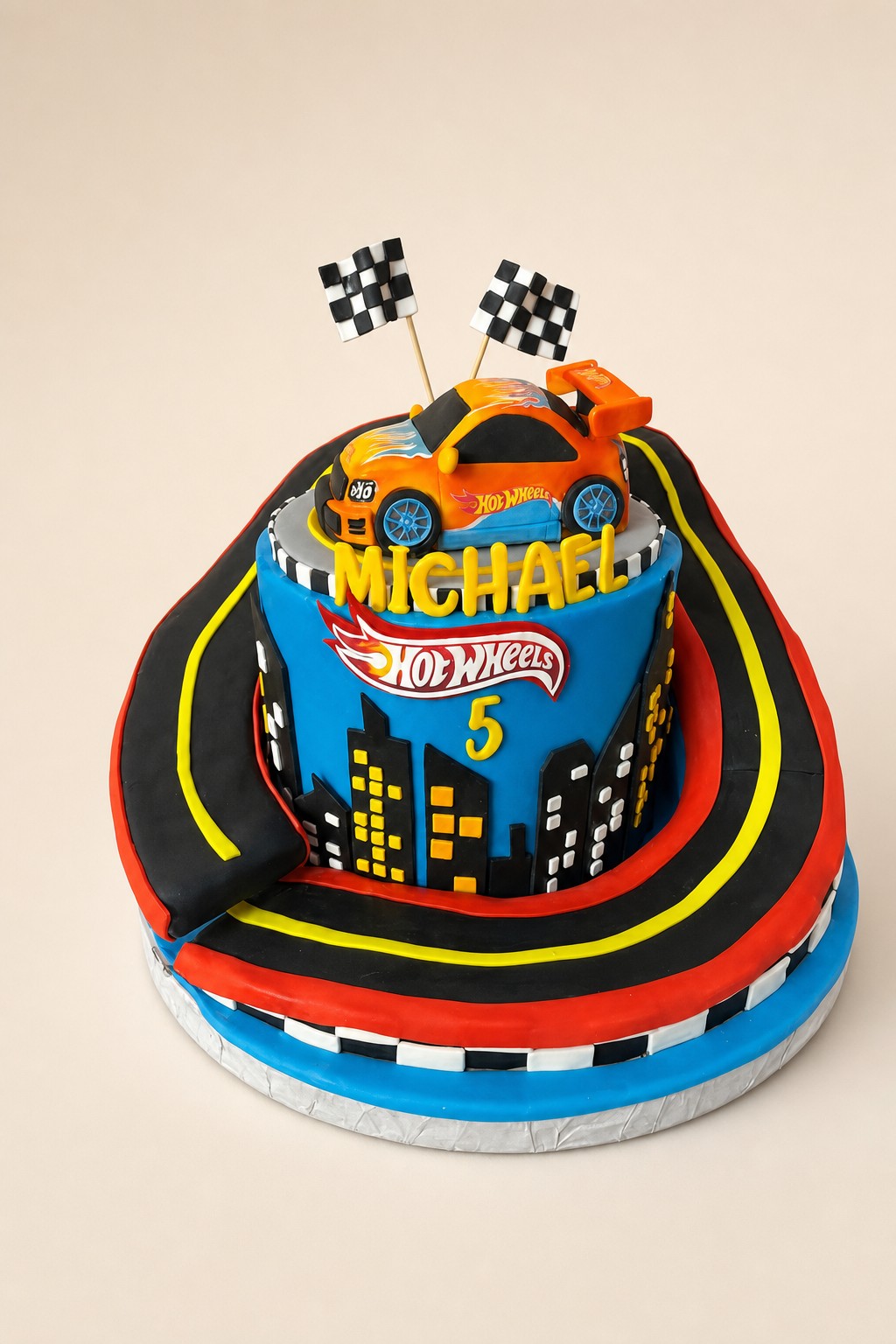 A vibrant Hot Wheels themed two-tier birthday cake for Michael's 5th birthday, featuring an orange race car, a city skyline, racing flags, a
