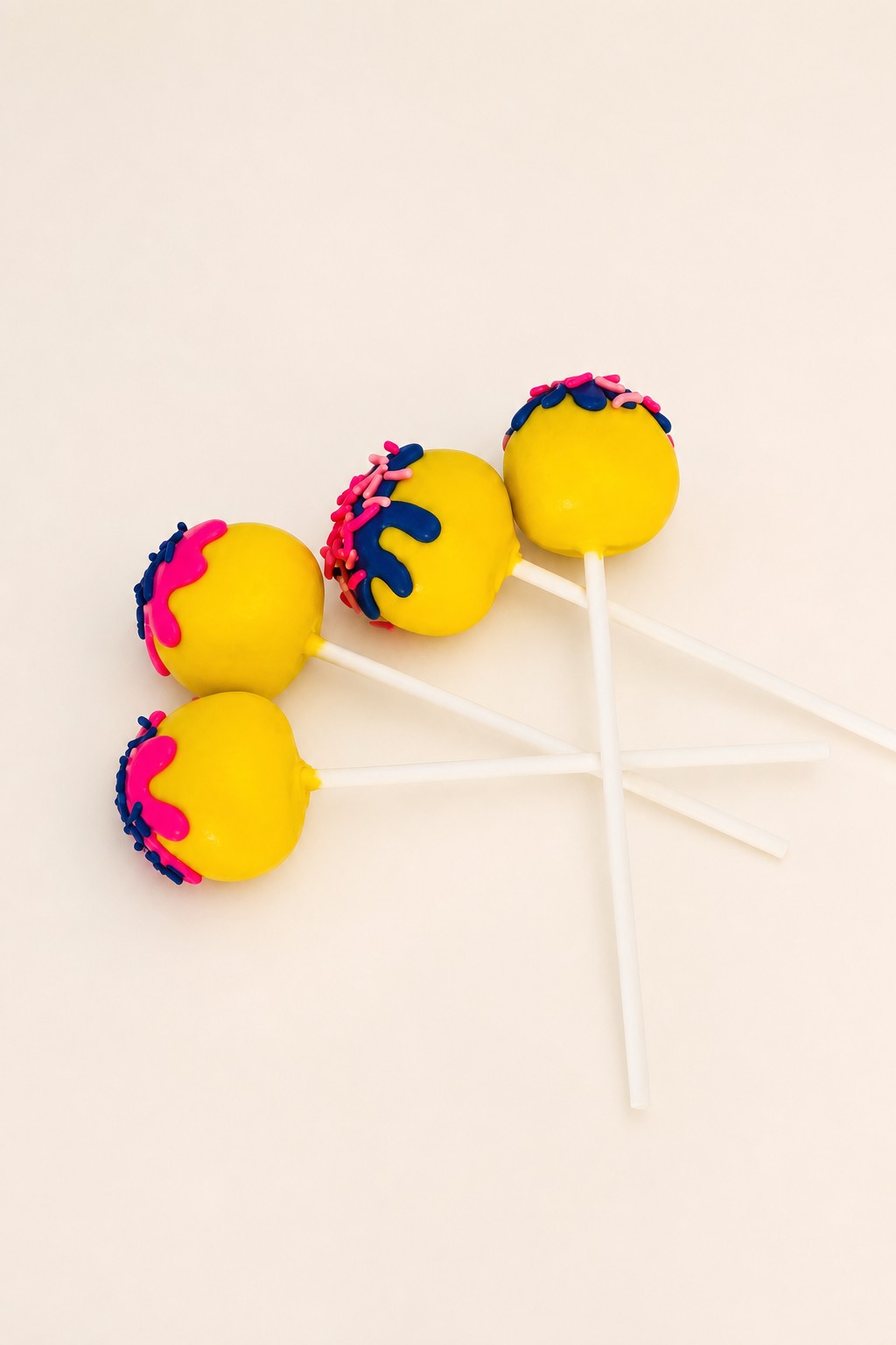 Four bright yellow cake pops decorated with vibrant pink and blue drizzle and matching sprinkles, displayed on a white rectangular plate.