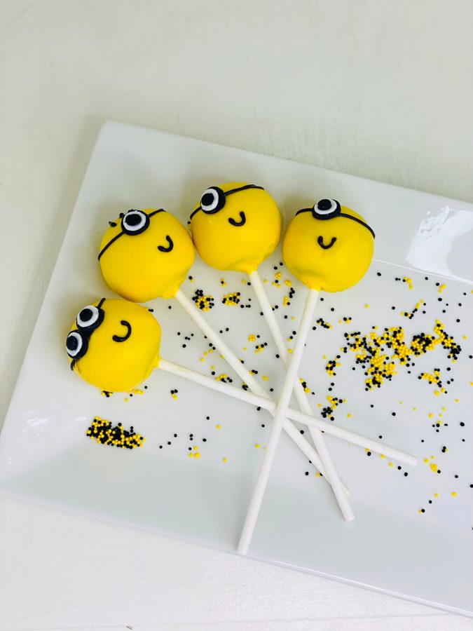 Minion-themed cake pops with yellow candy melt coating, black and white goggle decorations, and black smiles, presented on a white plate wit