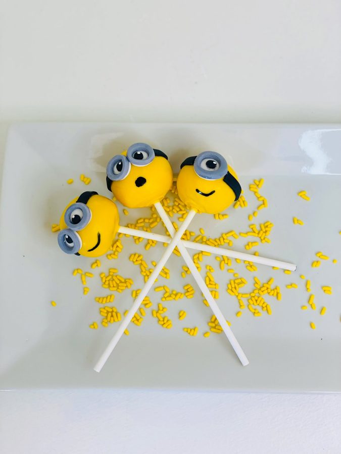 Three yellow Minion cake pops with grey goggles and black smiles, arranged on a white plate with scattered yellow sprinkles.