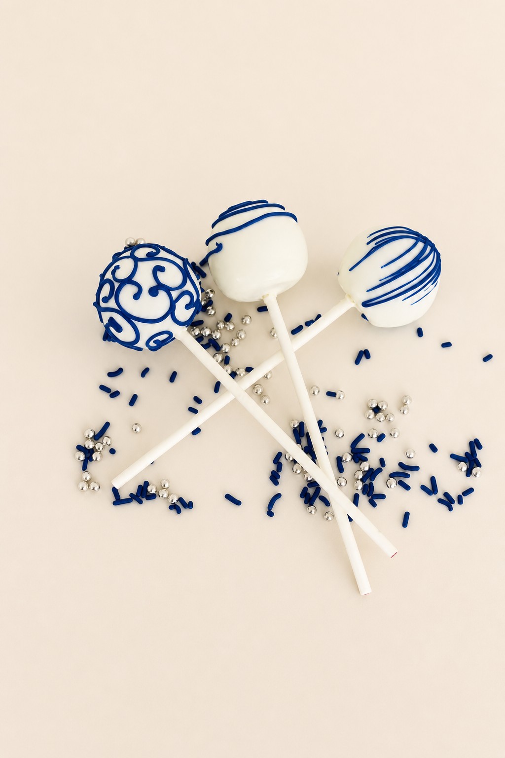 Three white cake pops, two decorated with blue stripes and one with elaborate blue swirl designs, resting on a white plate scattered with na