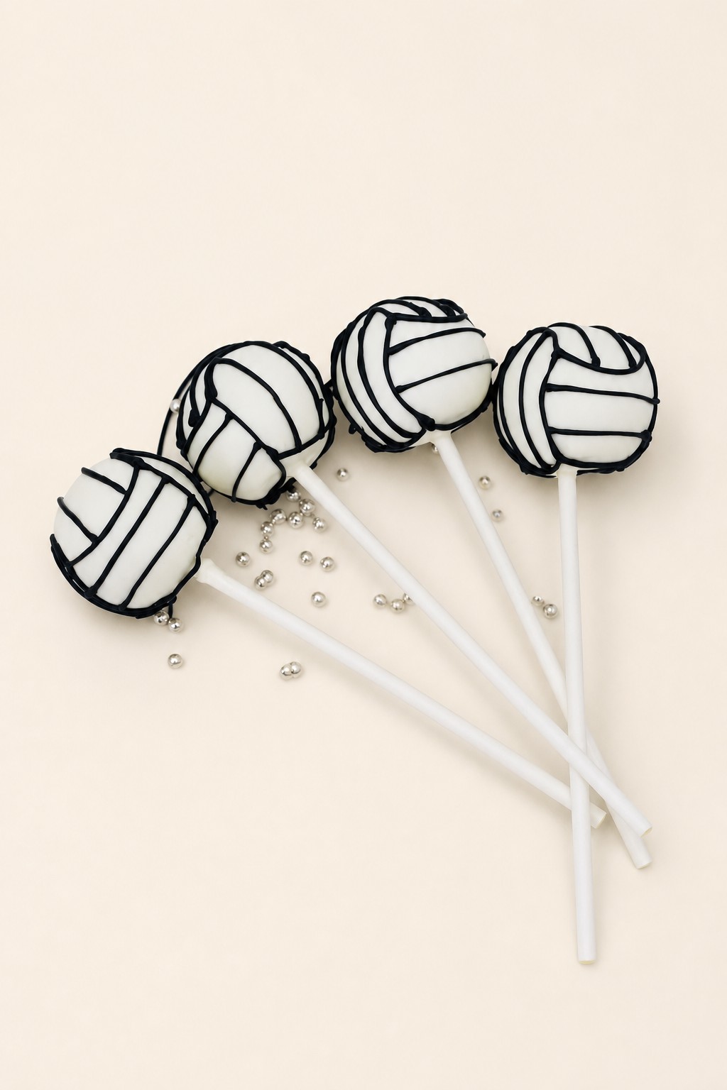 White cake pops decorated with black lines to resemble volleyballs, arranged on a white rectangular plate with scattered silver sprinkles.