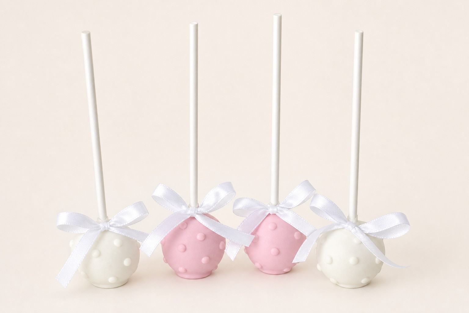 Four elegant cake pops, two white and two pink, each adorned with small raised dots and a delicate white ribbon bow, arranged on a light gre