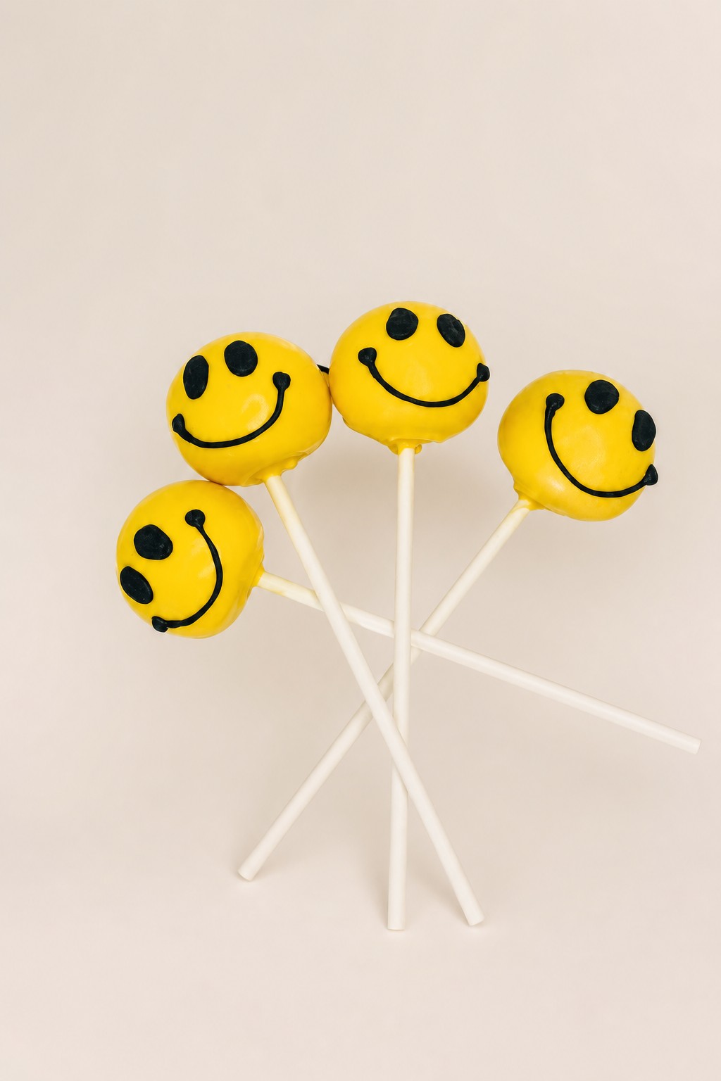 Four cheerful yellow smiley face cake pops with black icing details, presented on a white rectangular plate with yellow sprinkles.
