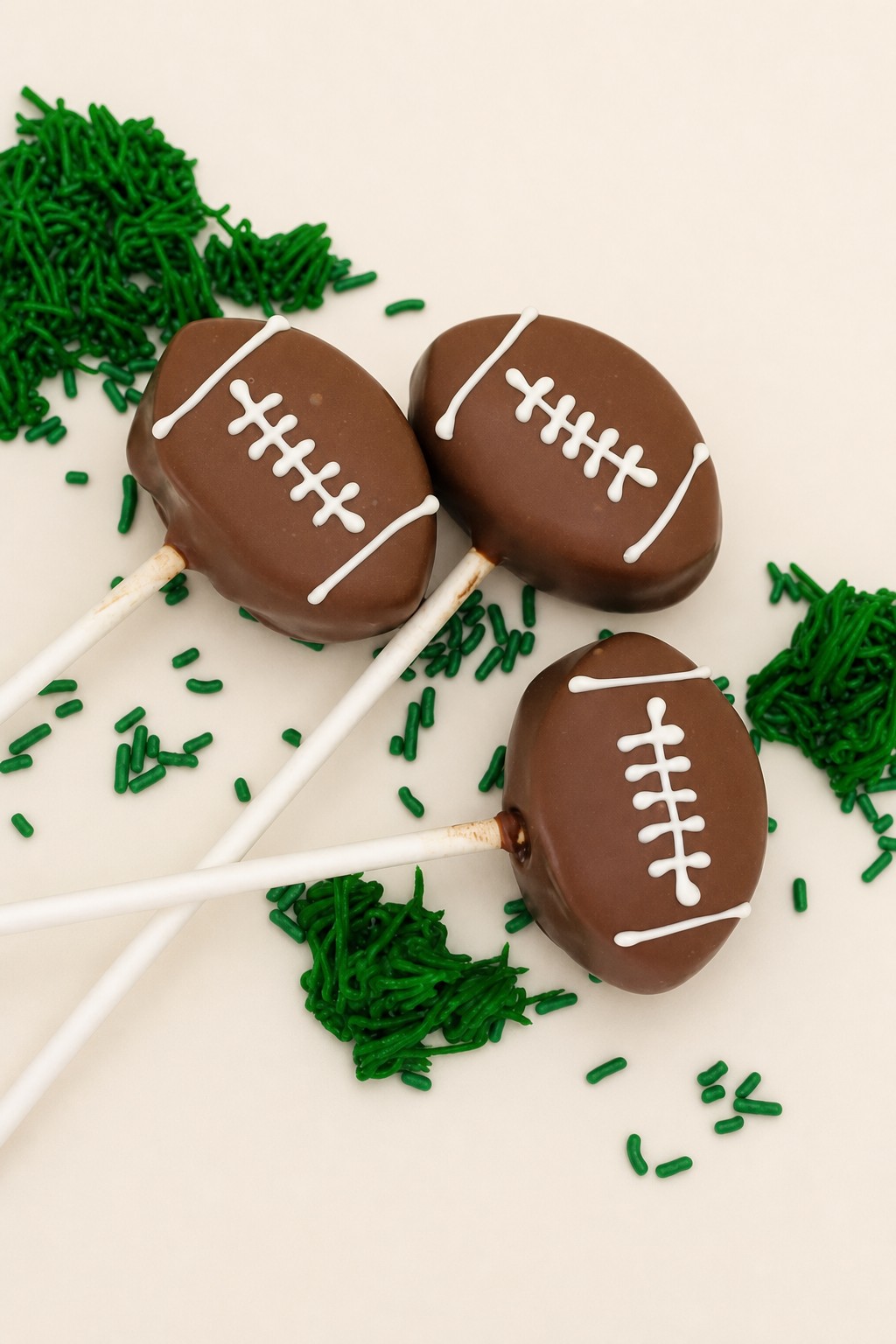 Three chocolate-covered football cake pops decorated with white icing laces and scattered green sprinkles and piped green icing resembling g