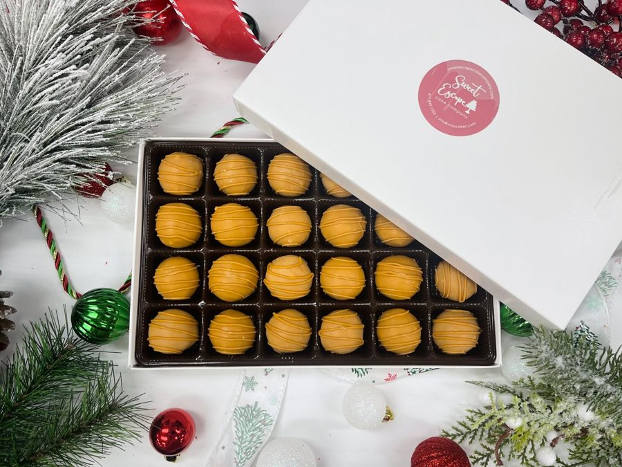 A box of festive round, orange-coated cake pops with caramel-colored drizzle, presented with Christmas ornaments and frosted pine needles.