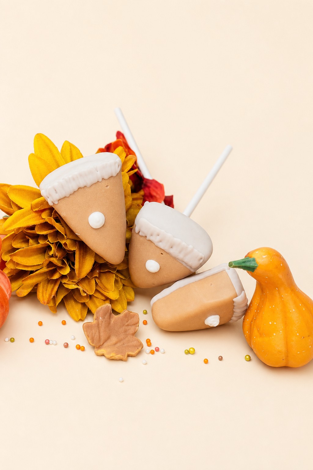 Autumn-themed cake pops or dessert treats shaped like pumpkin pie slices and acorns, decorated with cream-colored icing and sprinkles, prese