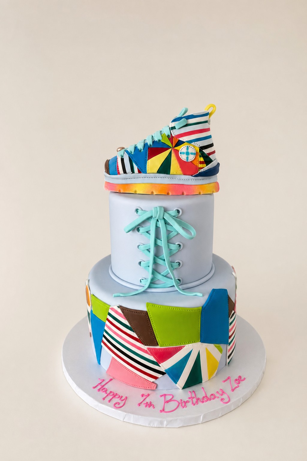 A vibrant two-tier round birthday cake featuring a colorful patchwork design, a detailed sneaker figurine on top, and light blue fondant lac