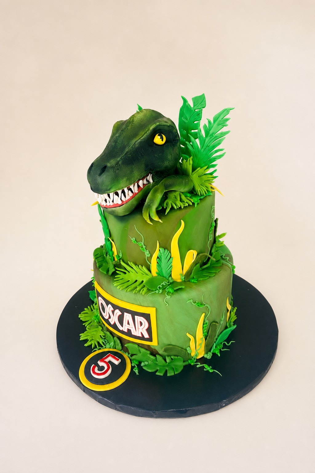 A two-tiered green fondant dinosaur birthday cake featuring a sculpted dinosaur head, jungle leaves, and personalized with the name OSCAR an