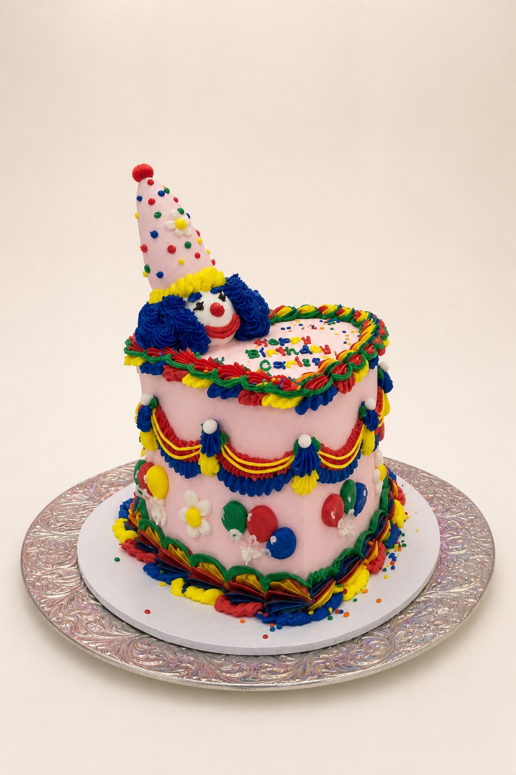 A vibrant pink buttercream birthday cake featuring a cheerful clown topper with a polka-dot party hat, adorned with colorful piped garlands,