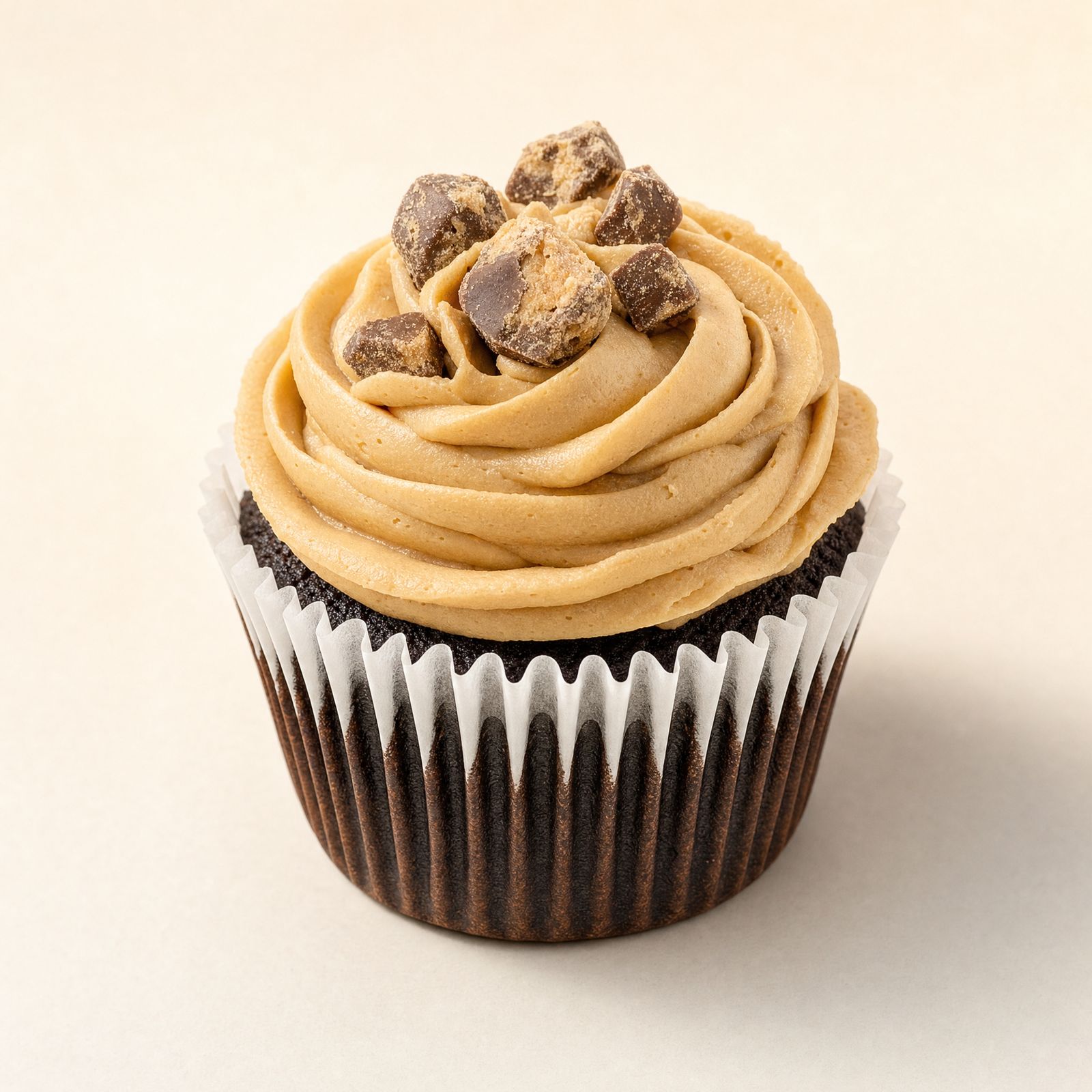 Two chocolate cupcakes with tan-colored buttercream frosting, topped with crunchy brown candy pieces, presented on a reflective gold display