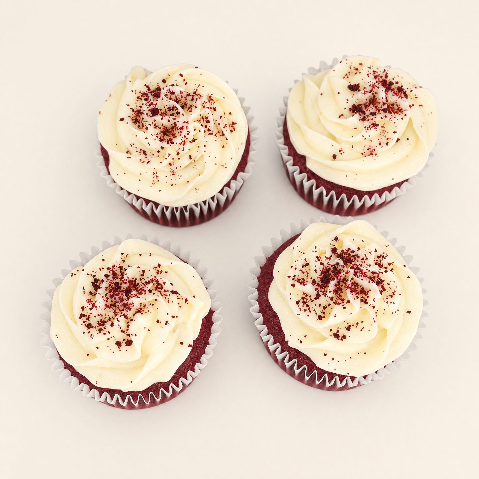 Four beautifully decorated red velvet cupcakes with creamy white frosting and red crumb sprinkles, presented on a minimalist white backgroun