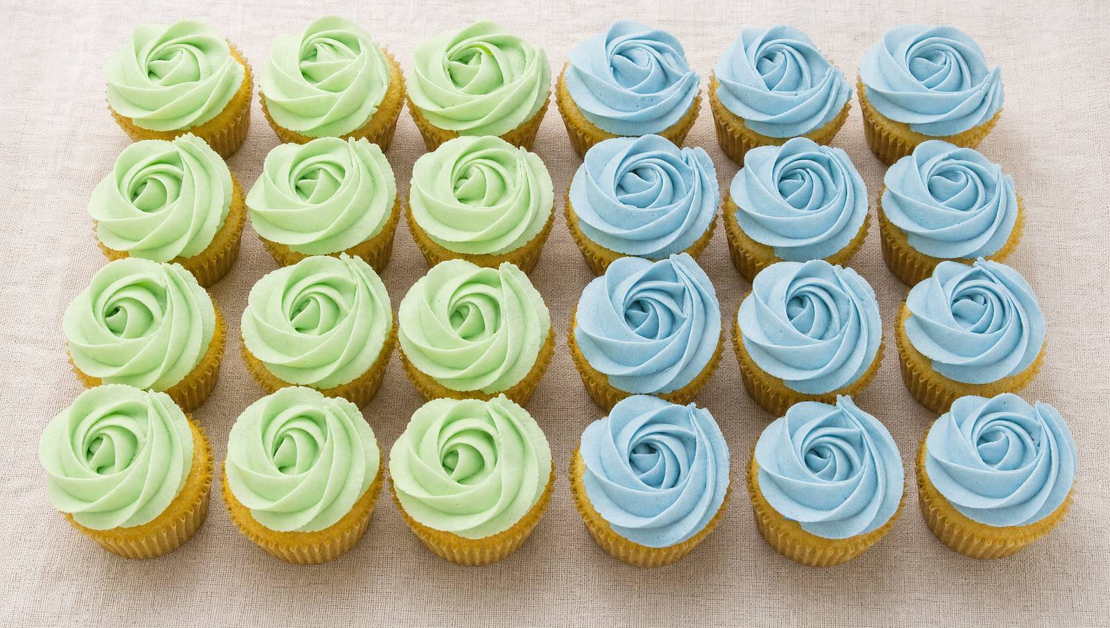 A clear tray of cupcakes featuring light green and light blue rose-shaped buttercream frosting, perfect for a baby shower or gender reveal c