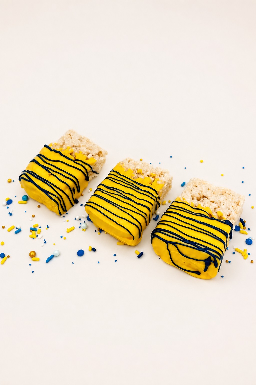 Three rectangular Rice Krispie treats partially dipped in bright yellow candy coating, decorated with thin blue stripes, and surrounded by c