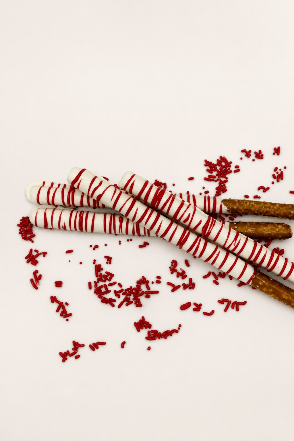 White chocolate-dipped pretzel rods with festive red stripes and scattered red sprinkles, presented on a white serving platter.