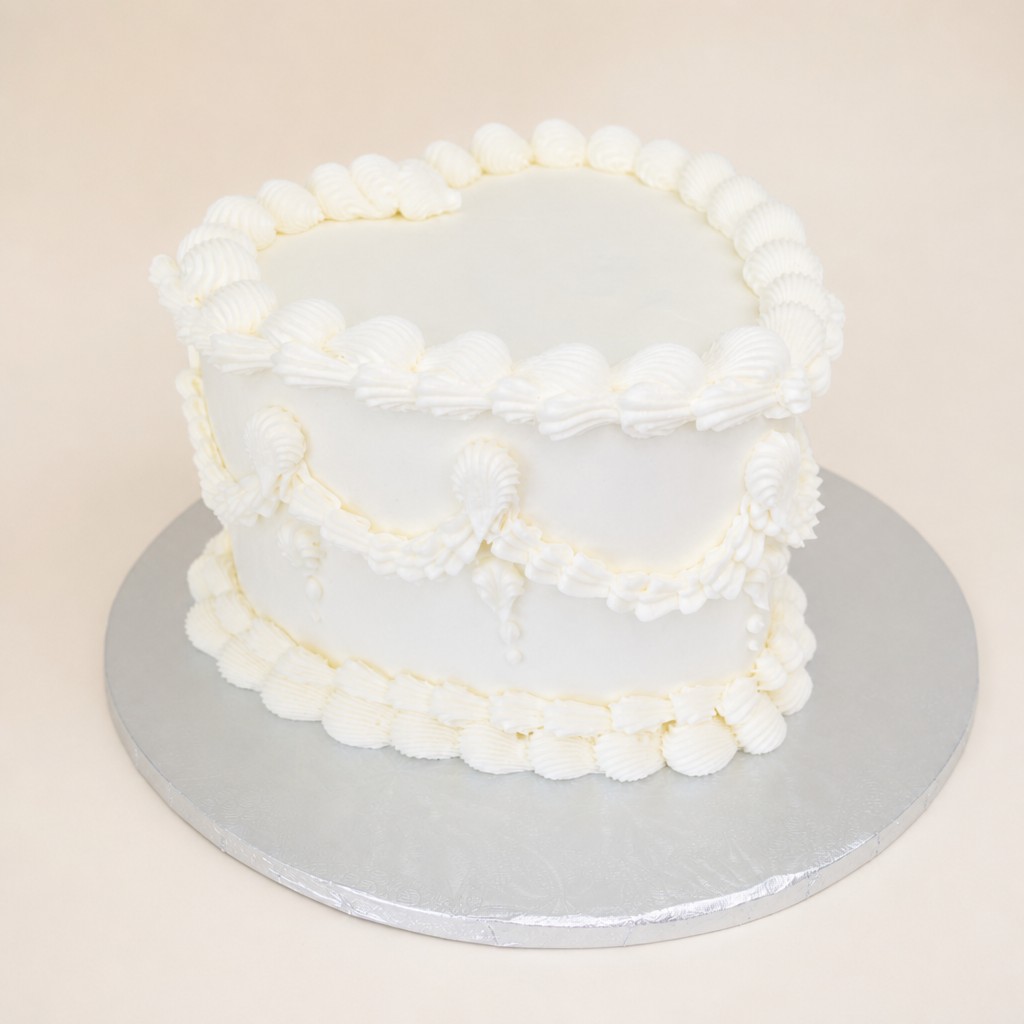 An elegant white heart-shaped cake adorned with intricate buttercream piping, featuring shell borders on the top and bottom edges, and ruffl