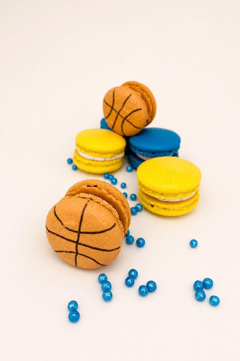 Custom basketball themed orange macarons, alongside yellow and blue macarons, decorated with blue sprinkles on a white platter.
