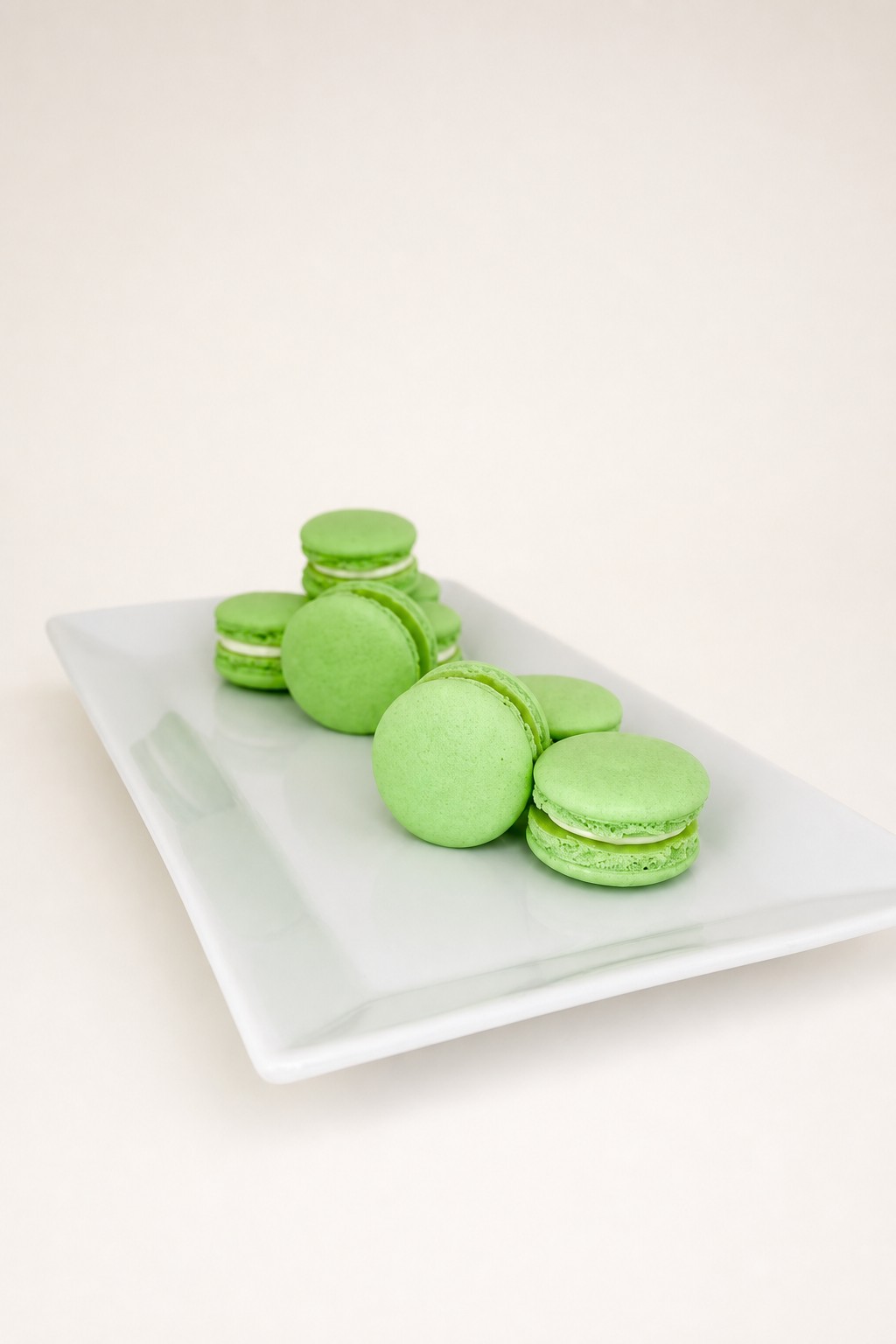 A collection of light green macarons with white filling is neatly arranged on a white rectangular serving plate, presented against a clean w