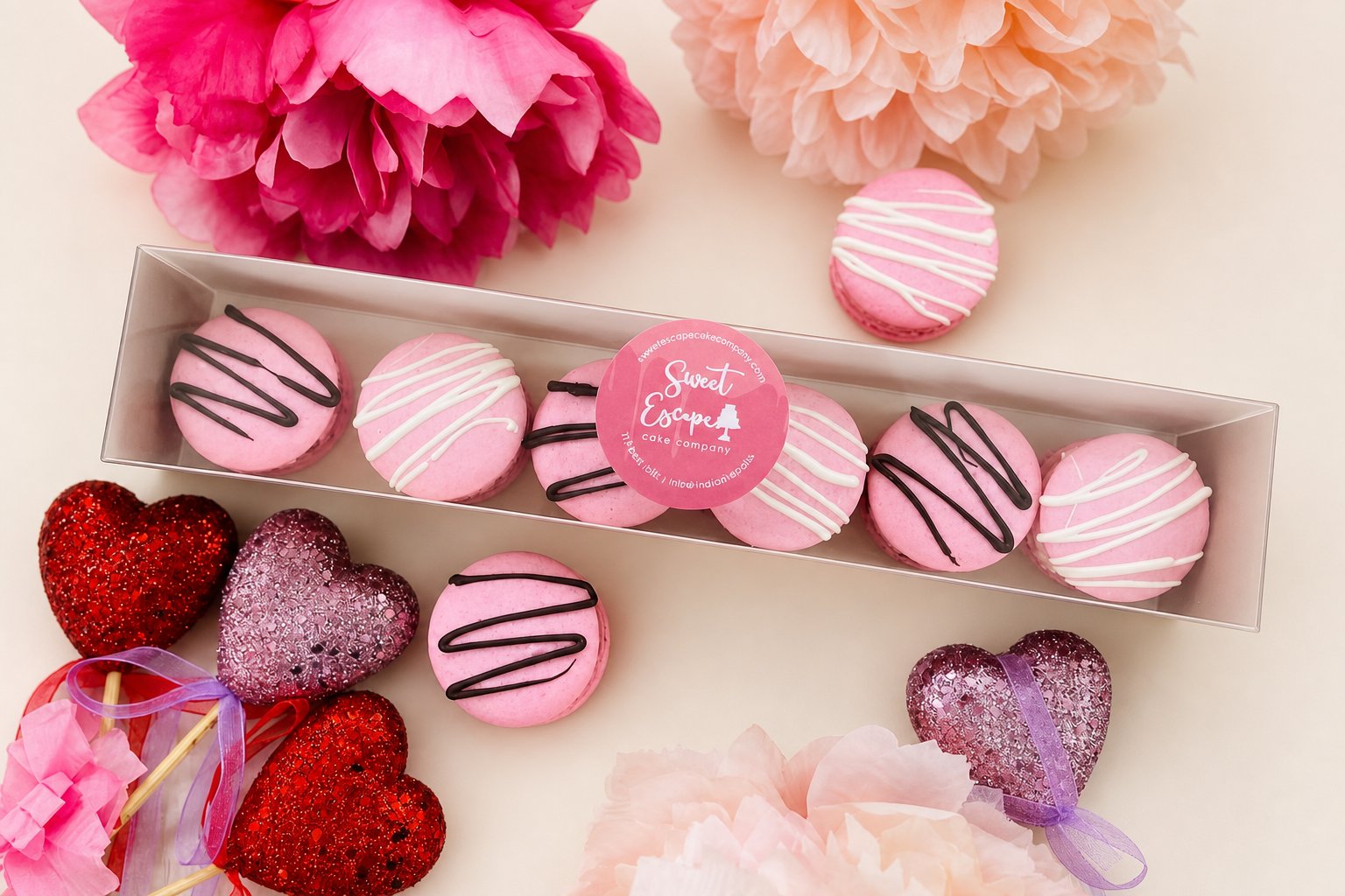 A box of round pink macarons, some decorated with white drizzle and others with black drizzle, presented in a festive setting with pink and 