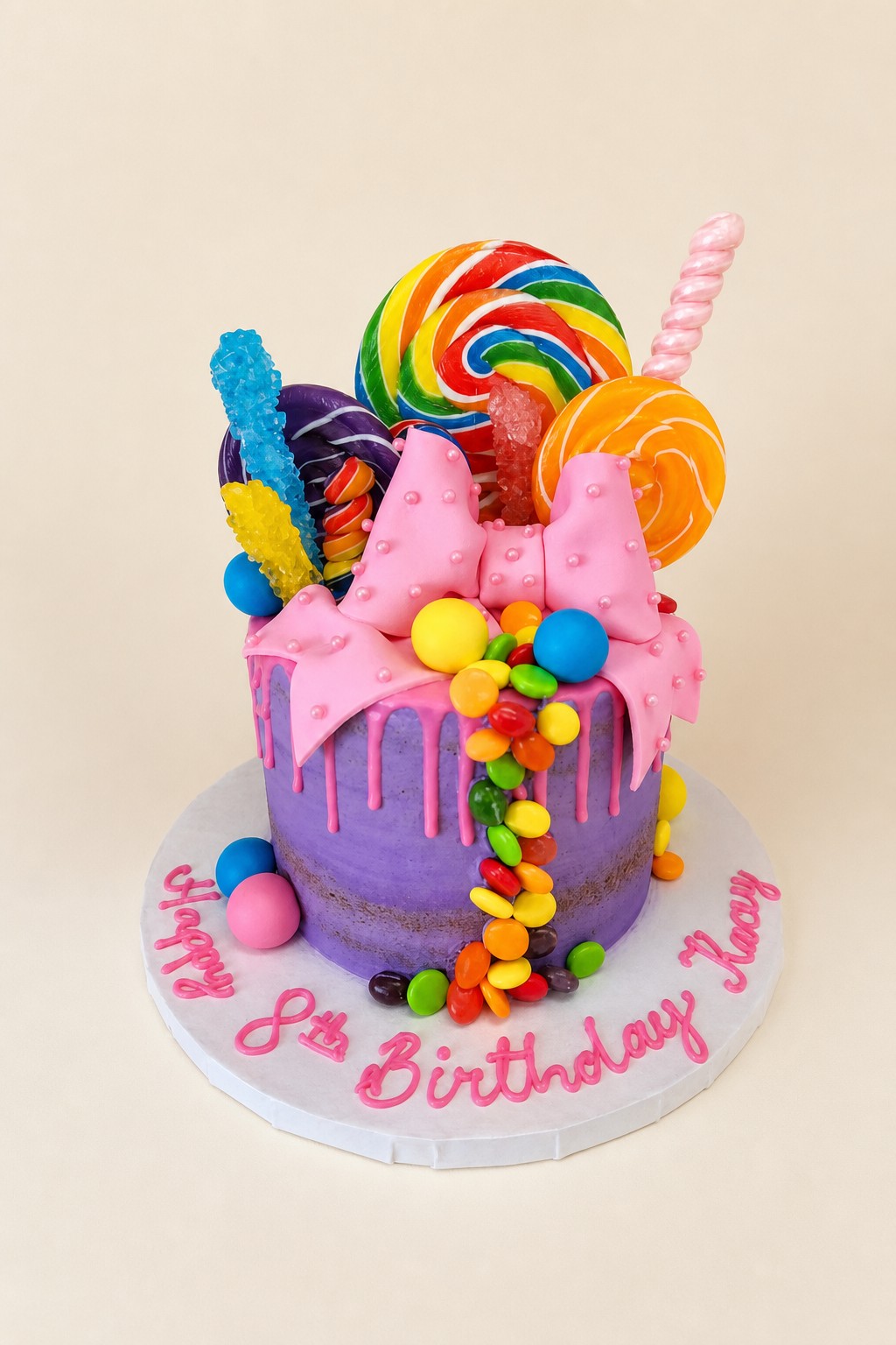 A vibrant single-tier purple drip cake adorned with a pink fondant bow, various lollipops, rock candy, and colorful candies, featuring "Happ